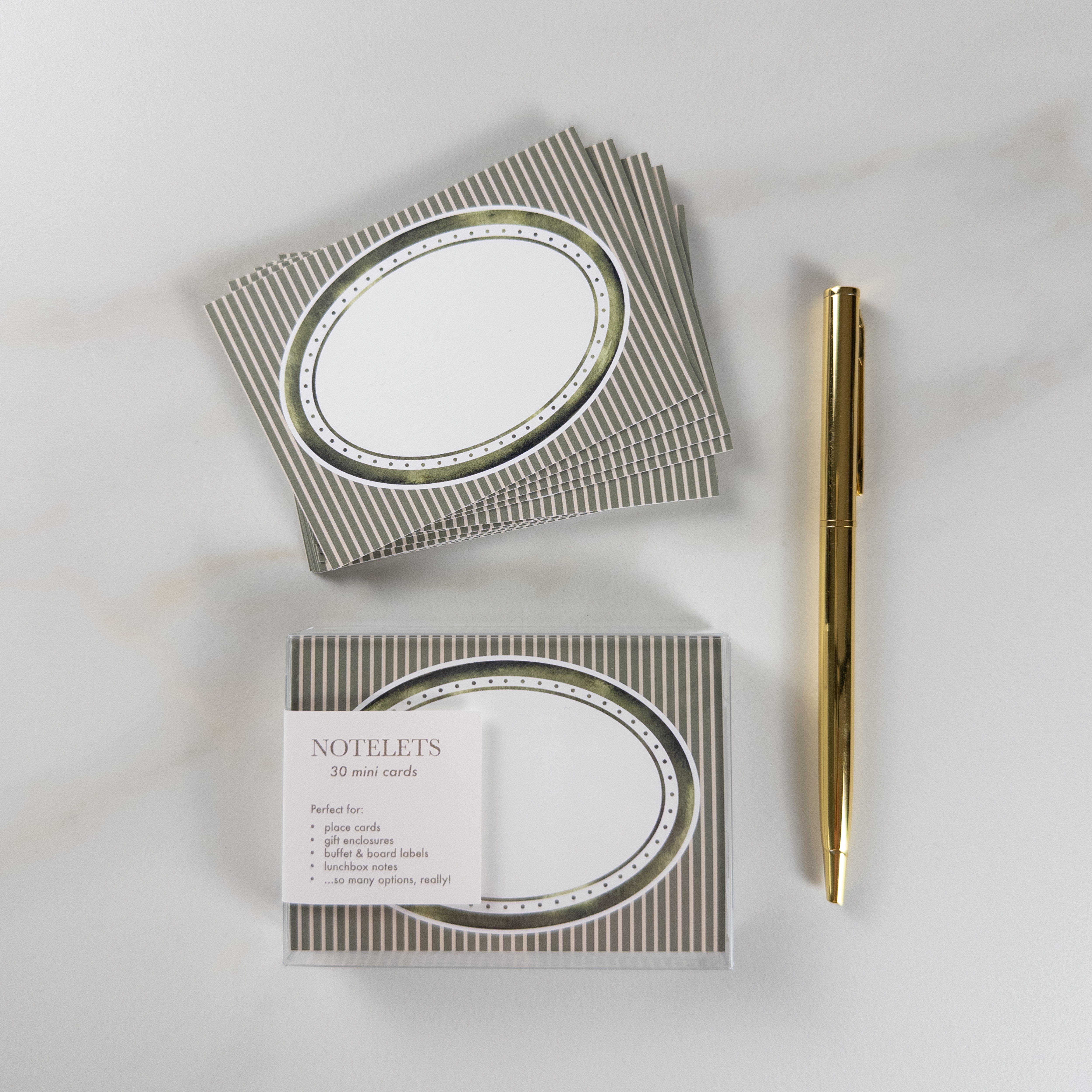 A set of oval notepads with metallic gold and silver striped covers, shown with one open pad and one closed pack labeled 'NOTELETS', alongside a gold pen on a light marble background.